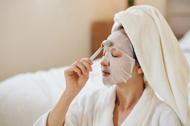 Mulher desfrutando de um momento de autocuidado, massageando o rosto com um rolo de jade enquanto usa uma máscara de folha iluminadora para realçar a beleza natural da pele.